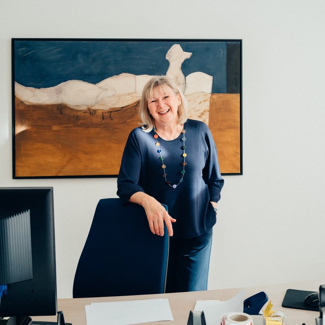 Eine Frau steht in einem Büro und lächelt, hinter ihr hängt ein Gemälde an der Wand.