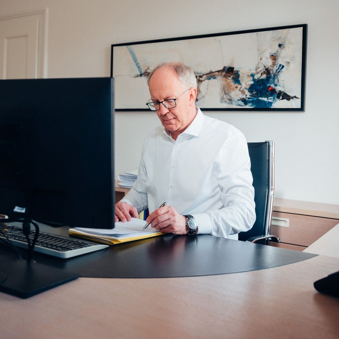 Ein älterer Mann in einem weißen Hemd sitzt an einem Schreibtisch und schreibt in ein Notizbuch, ein Computerbildschirm steht vor ihm.