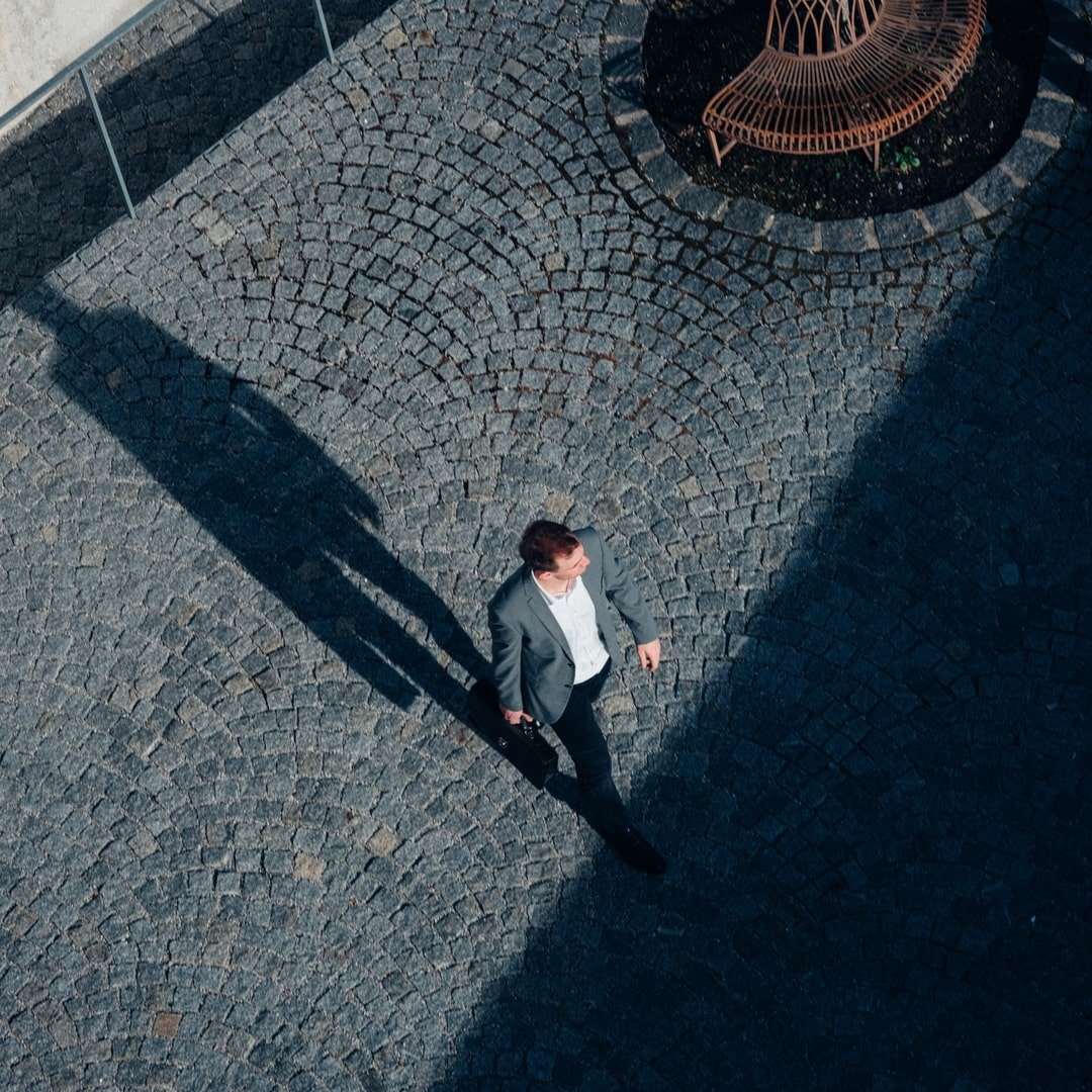 Luftaufnahme eines Mannes im Anzug, der über einen gepflasterten Platz geht, der Schatten fällt auf den Boden.