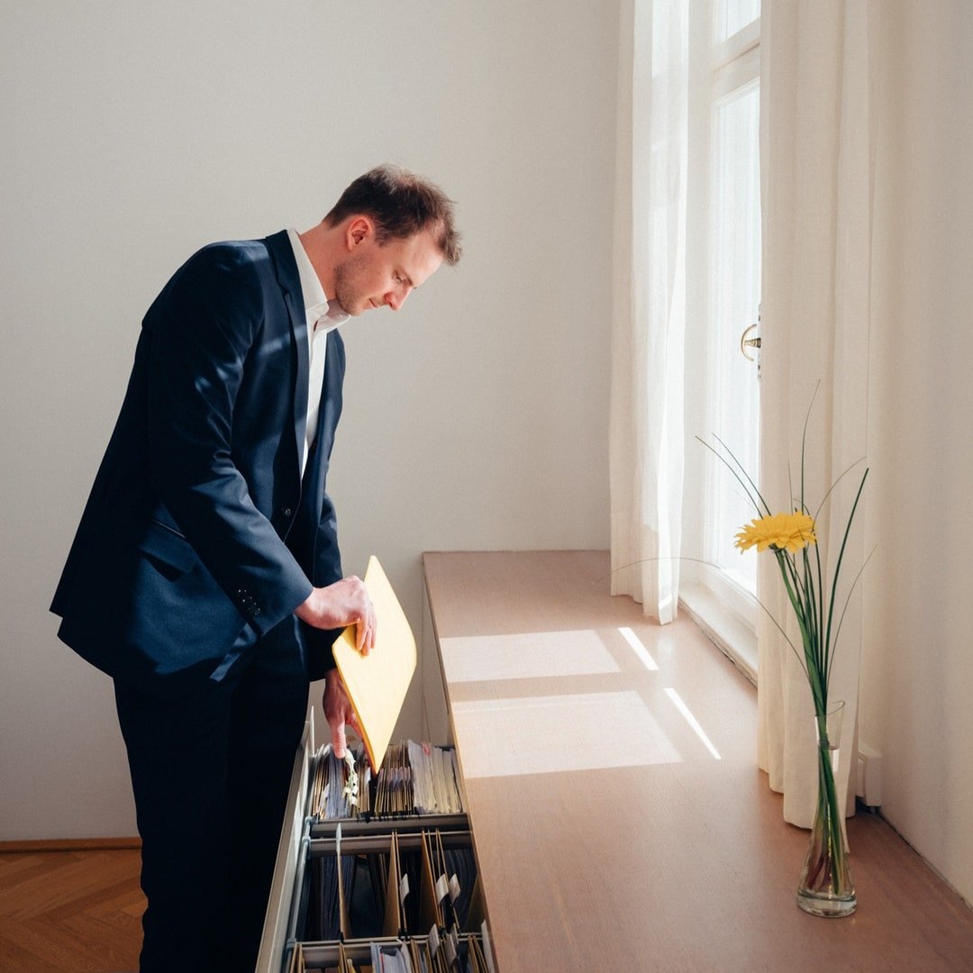 Ein Mann in einem Anzug durchsucht Akten in einem Aktenschrank neben einem Fenster.