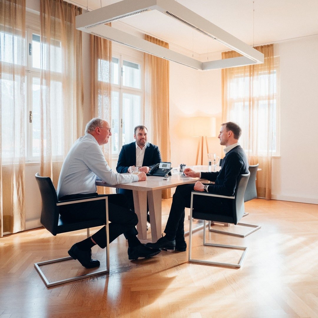 Weitwinkelaufnahme eines Meetings mit drei Männern, die um einen Tisch sitzen und diskutieren.