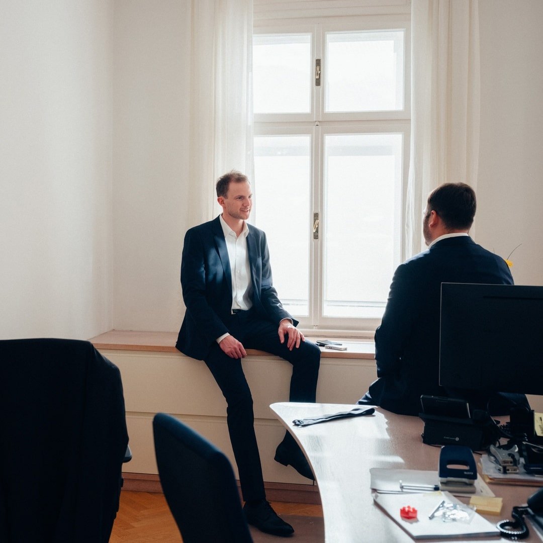 Zwei Männer in einem Büro, einer sitzt auf der Fensterbank, der andere auf einem Stuhl, beide in einem Gespräch vertieft.